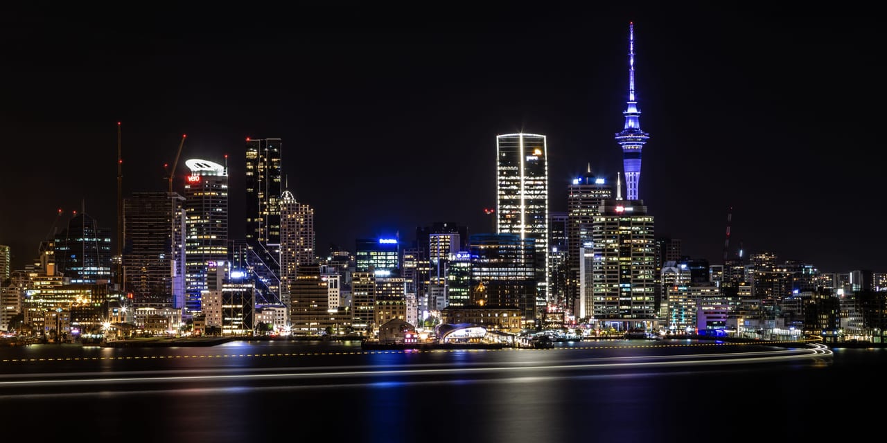A wide view of Auckland's skyline illuminated at night with the Sky Tower visible.